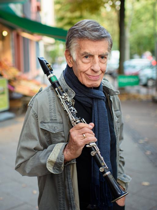 Rolf Kühn, Jazzmusiker und Komponist, steht im Berliner Bezirk Charlottenburg auf einem Gehweg und hält seine Klarinette in der Hand. Er hat graue, zur Seite gekämmte Haare und trägt einen blauen Schal um den Hals.