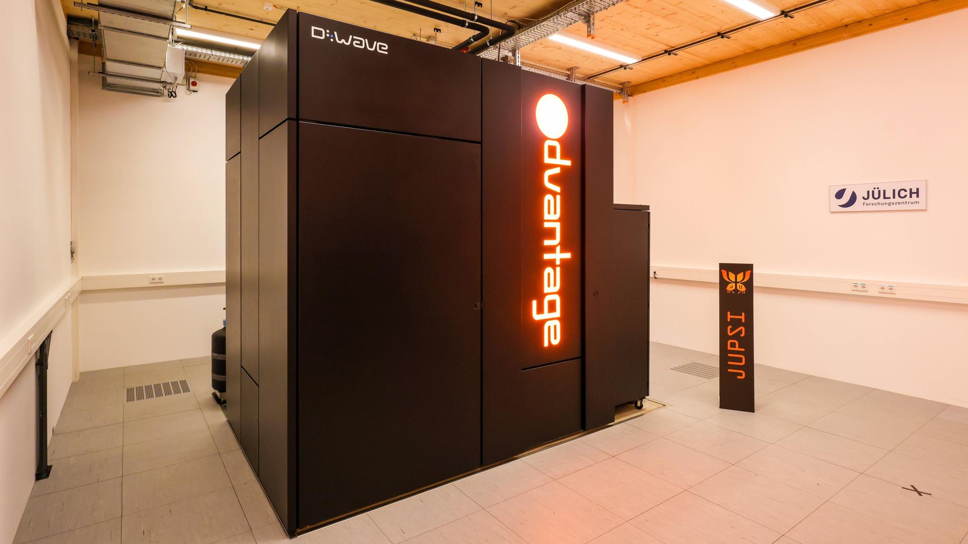 Ein schwarzer Quantencomputer steht in einem Raum mit Holzdecke im Forschungszentrum Jülich. Auf dem Computer steht in roten Leuchtbuchstaben "Advantage", der Name des Rechners.