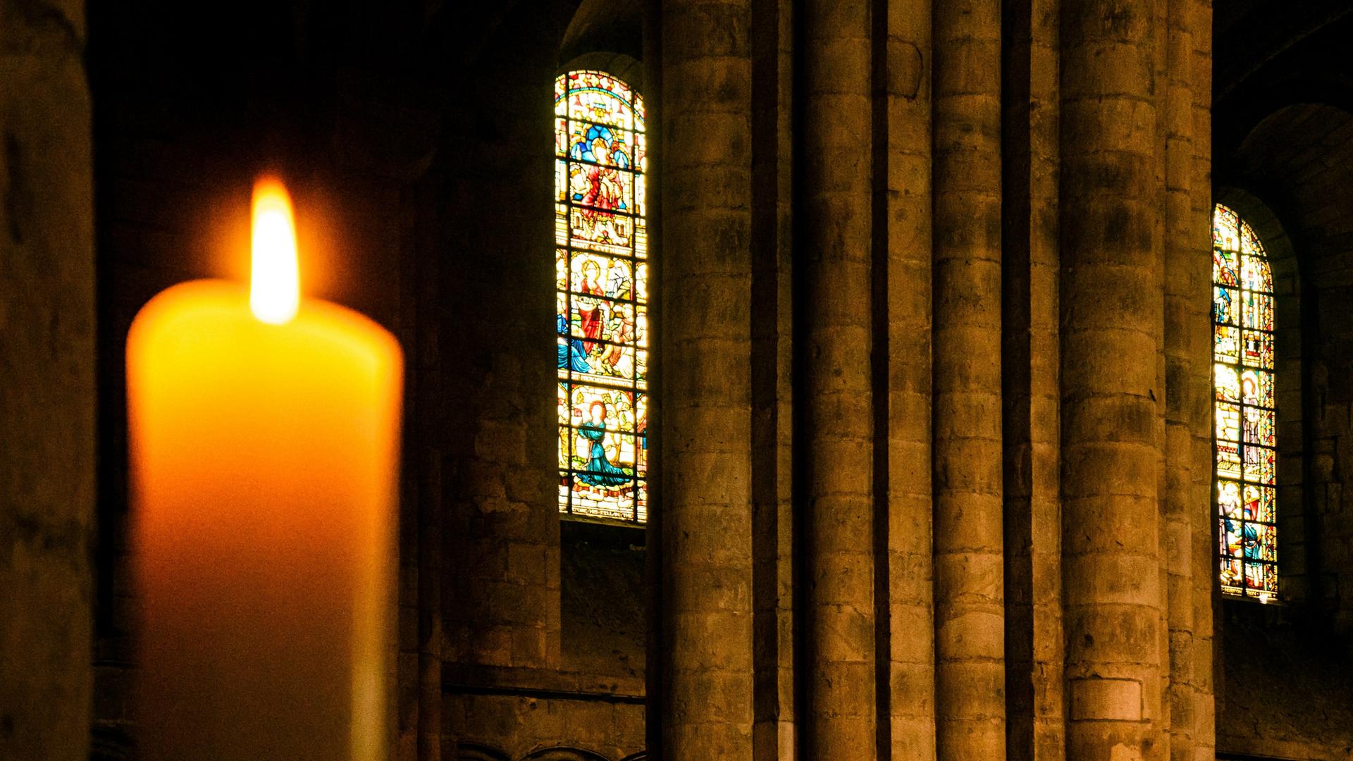 Blick auf eine Kerze im Vordergrund, die in einem Kirchenraum brennt. Im Hintergrund erkennt man ein buntes Glasfenster,.