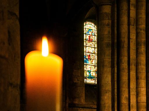 Blick auf eine Kerze im Vordergrund, die in einem Kirchenraum brennt. Im Hintergrund erkennt man ein buntes Glasfenster,.