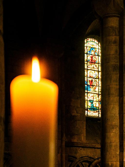 Blick auf eine Kerze im Vordergrund, die in einem Kirchenraum brennt. Im Hintergrund erkennt man ein buntes Glasfenster,.