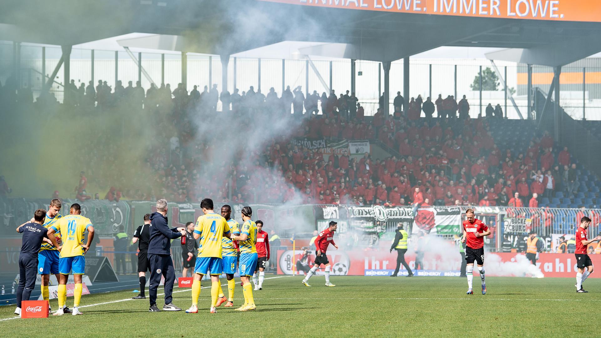 Mehrere Fußballspieler warten auf dem Spielfeld, während im Hintergrund Pyrotechnik brennt. Mehrere Fußballspieler warten auf dem Spielfeld, während im Hintergrund Pyrotechnik brennt.