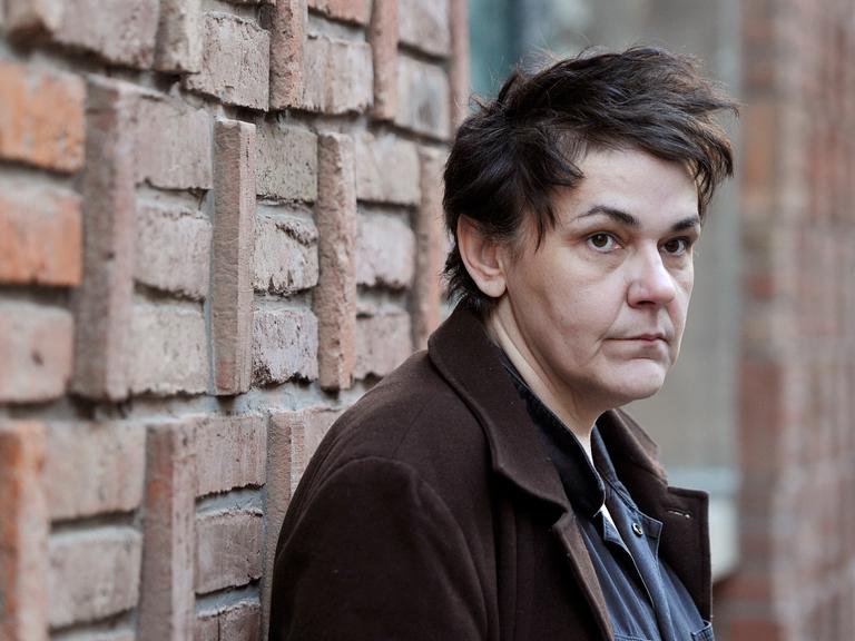 Die Sängerin Christiane Rösinge steht vor einer Wand und blickt in Richtung Kamera. Sie hat verwuscheltes Haar und trägt einen Mantel.
