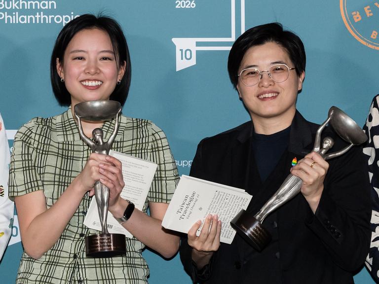 Zwei Frauen stehen nebeneinander und lächeln. Sie halten jeweils eine Statuette in silber.