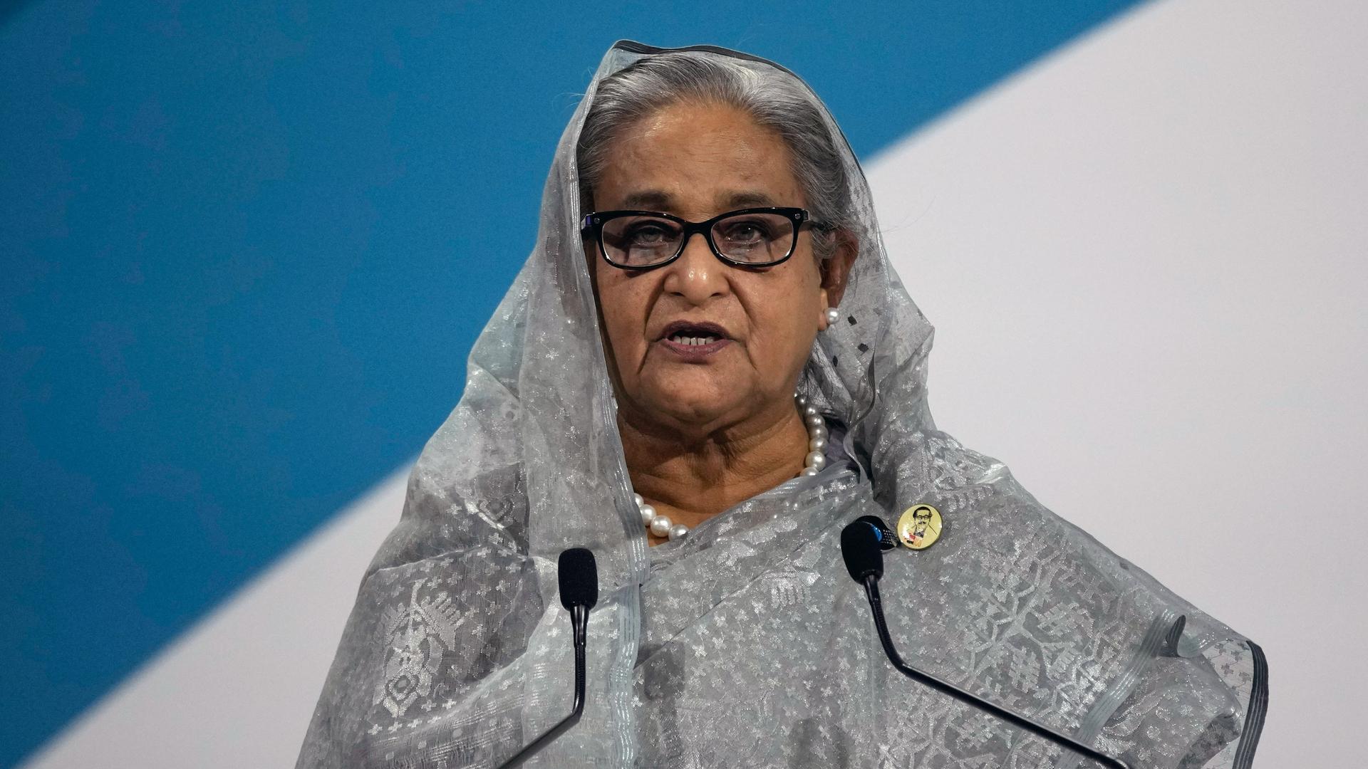 Hasina mit Kopfftuch und grauem Kleid steht vor einer halb blauen, halb weißen Wand und spricht in zwei Mikrofone. 