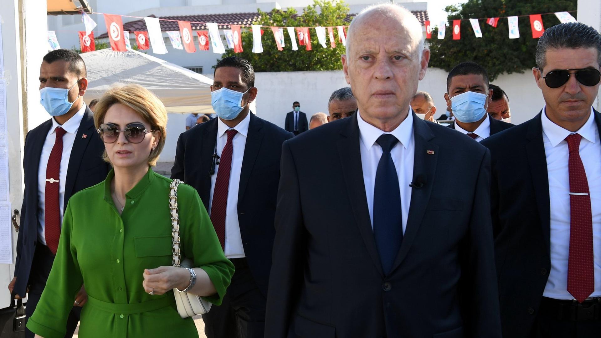 Tunesiens Präsident Kais Saied, begleitet von seiner Ehefrau und mehreren Leibwächtern, geht durch eine mit Wimpeln geschmückte Straße auf die Kamera zu.