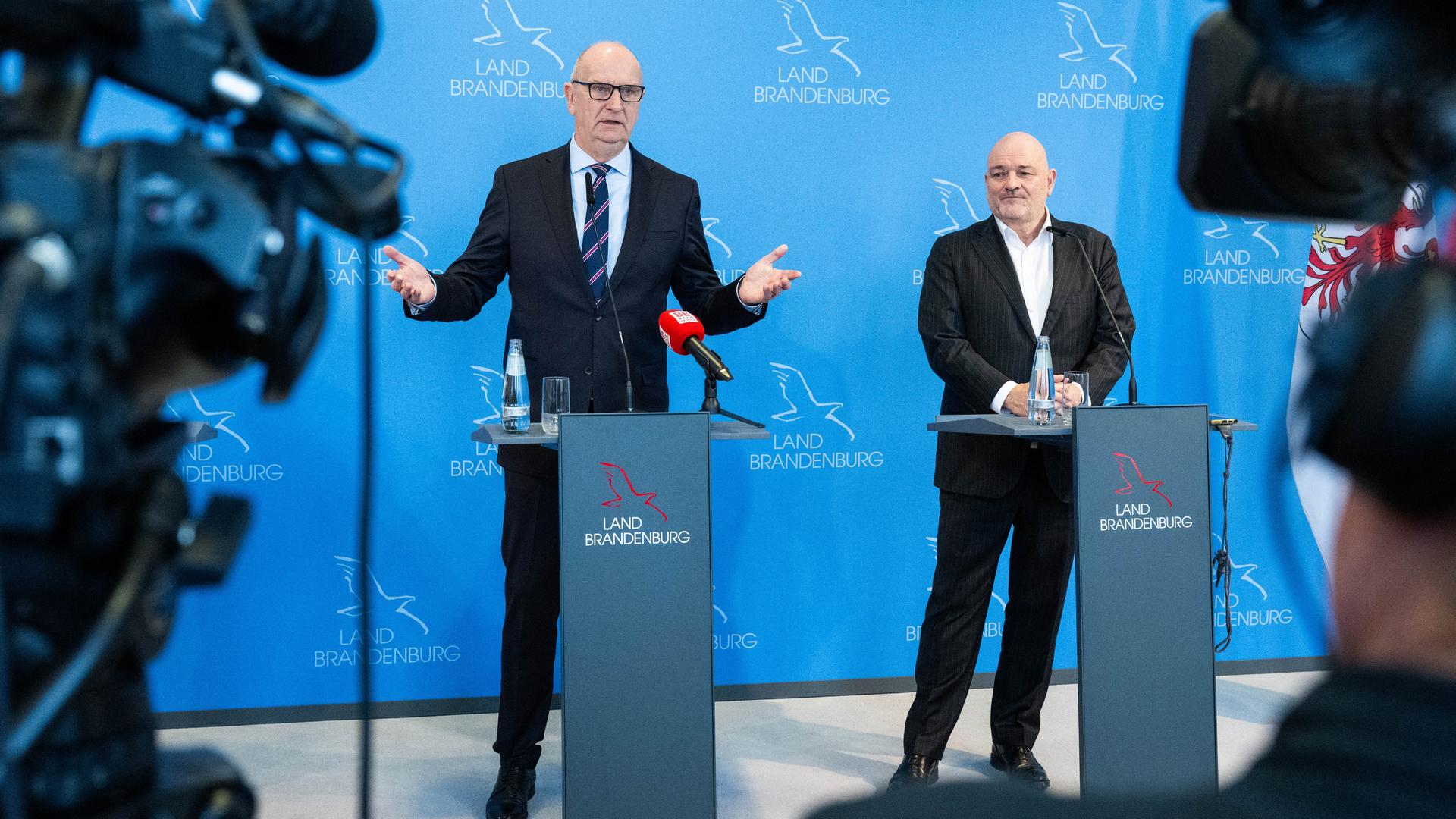 Dietmar Woidke (l.) und Robert Crumbach stehen in der Staatskanzlei von Brandenburg bei einem Statement für die Presse vor einer blauen Wand zusammen.