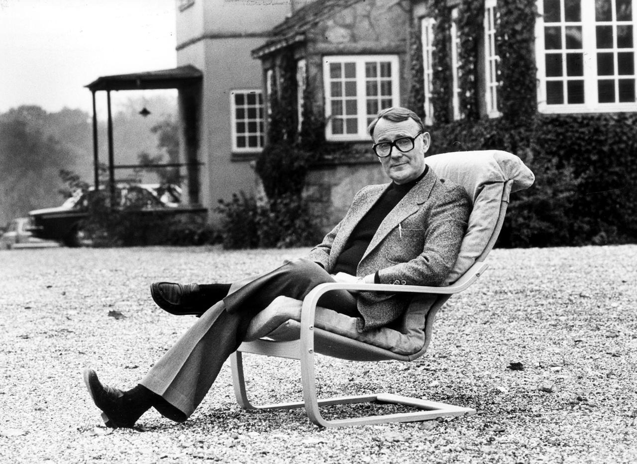 Ein älterer Mann sitzt auf einem Sessel, der im Garten vor einem Haus steht.
