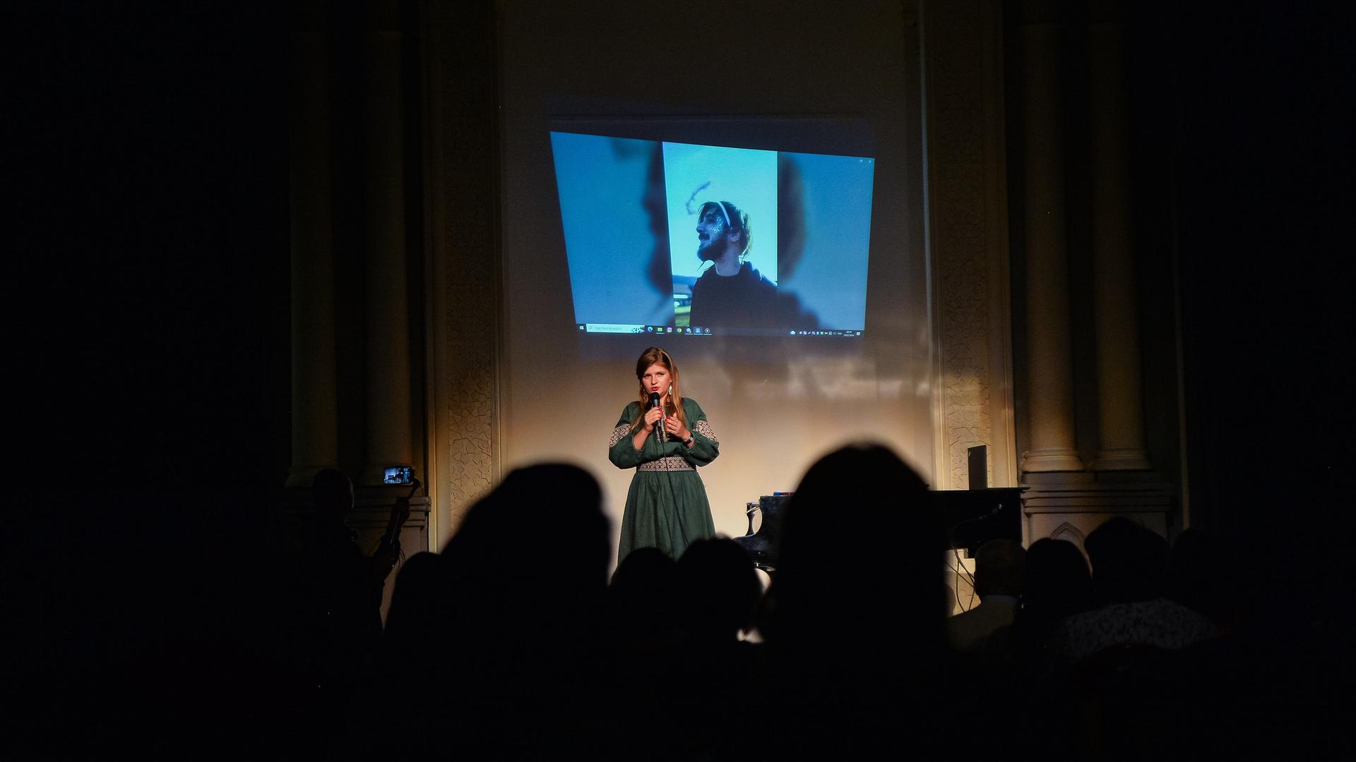 Bei einer Veranstaltung in Kiew wird an den im Krieg gefallenen Dichter Maksym Kryvtsov erinnert. Sein Porträt ist beleuchtet, im Vordergrund ist das Publikum im Dunkeln, eine Frau spricht. 
