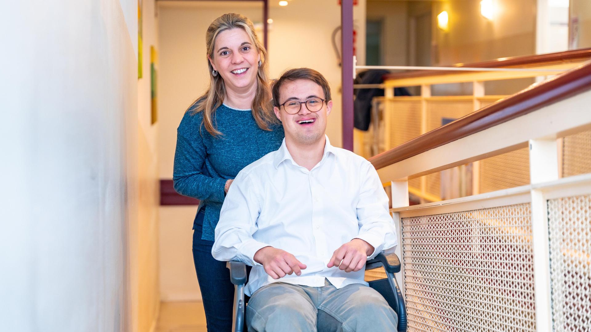 Eine Pflegerin steht hinter einem jungen Mann mit Down Syndrom, der lachend in einem Rollstuhl sitzt.