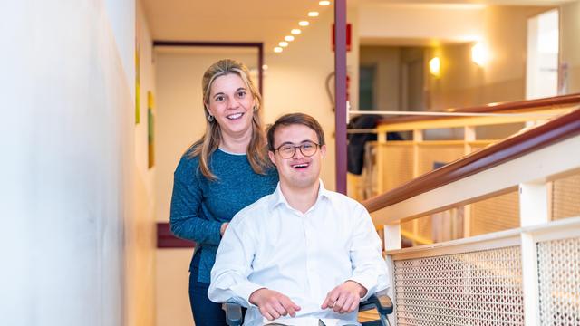 Eine Pflegerin steht hinter einem jungen Mann mit Down Syndrom, der lachend in einem Rollstuhl sitzt.