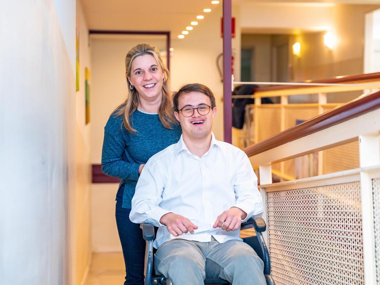 Eine Pflegerin steht hinter einem jungen Mann mit Down Syndrom, der lachend in einem Rollstuhl sitzt.