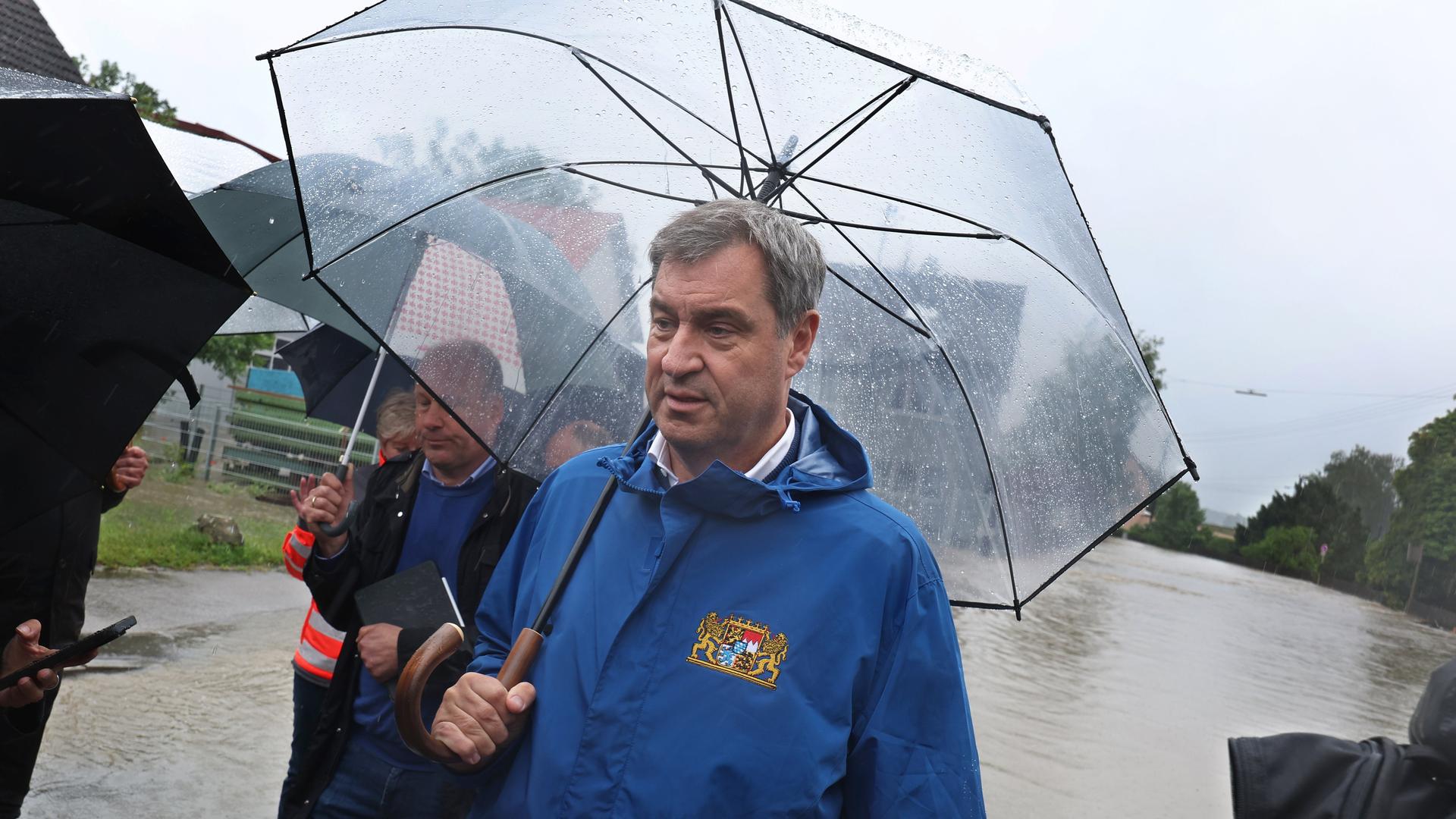 Markus Söder (CSU) steht am Rande einer überfluteten Straße in Diedorf und hält einen Regenschirm in der Hand.