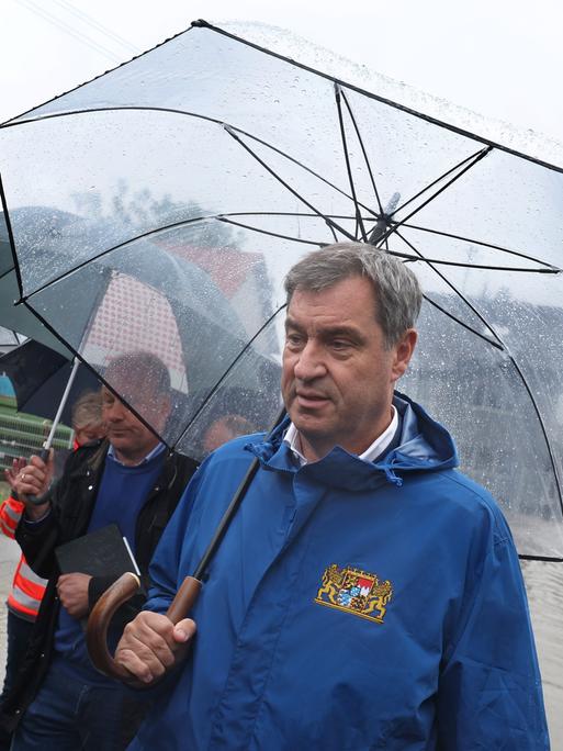 Markus Söder (CSU) steht am Rande einer überfluteten Straße in Diedorf und hält einen Regenschirm in der Hand.