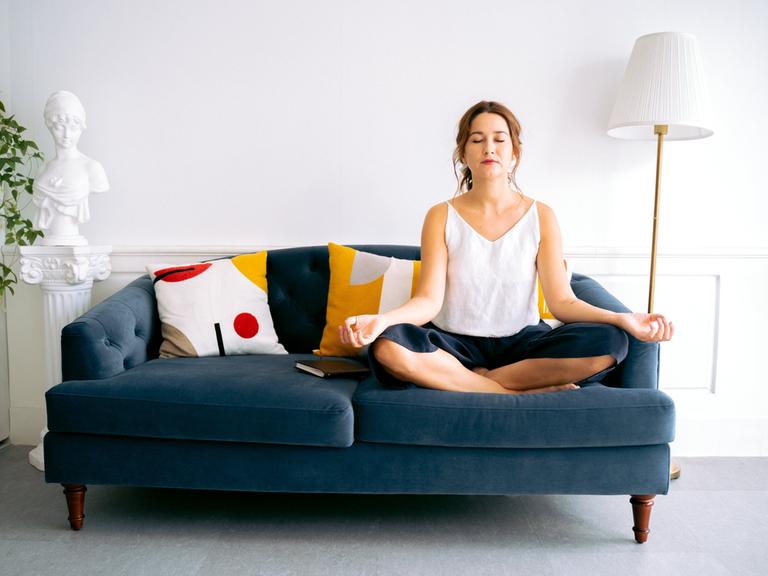 Eine Frau sitzt auf einem Sofa, hat ihre Augen geschlossen und meditiert. 