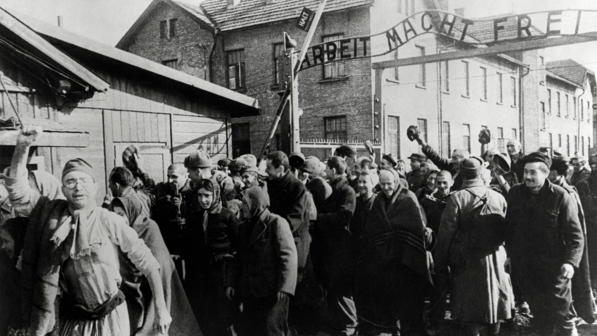 Schwarz-Weiß-Bild: Eine Menge Häftlingen tritt durch das Tor, über dem "Arbeit macht frei" steht. Manche strecken jubeldn die Arme nach oben. Links eine Baracke.