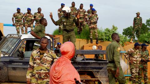 Nigerianische Polizei steht lächelnd auf einem Pick-Up-Truck während einer Demonstration von Putschisten vor dem französischen Stützpunkt in Niamey, Niger, am 11. August 2023. Im Vordergrund ist eine in orangenes Tuch gehüllte Passantin zu sehen. 