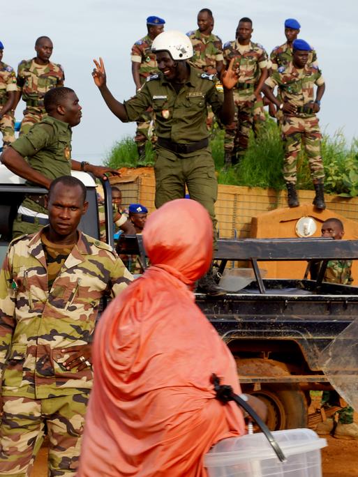 Nigerianische Polizei steht lächelnd auf einem Pick-Up-Truck während einer Demonstration von Putschisten vor dem französischen Stützpunkt in Niamey, Niger, am 11. August 2023. Im Vordergrund ist eine in orangenes Tuch gehüllte Passantin zu sehen. 