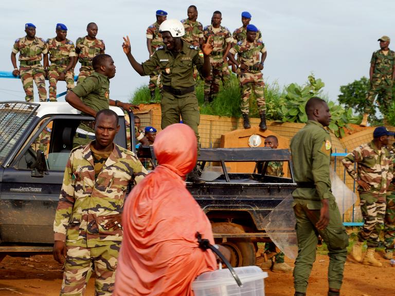 Nigerianische Polizei steht lächelnd auf einem Pick-Up-Truck während einer Demonstration von Putschisten vor dem französischen Stützpunkt in Niamey, Niger, am 11. August 2023. Im Vordergrund ist eine in orangenes Tuch gehüllte Passantin zu sehen. 