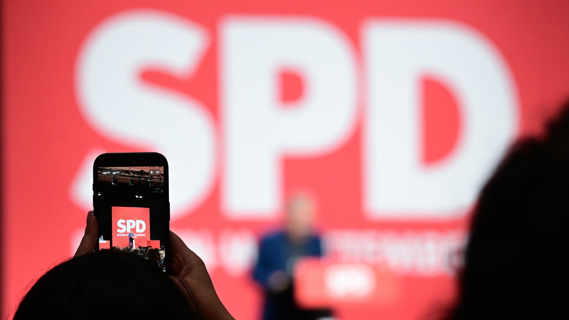 Auf einem der SPD-Parteitag filmt eine Delegierte den Redner am Rednerpult, den man im Hingtergrund unscharf unter einem großen SPD-Logo sprechen sieht. Auf einem der SPD-Parteitag filmt eine Delegierte den Redner am Rednerpult, den man im Hingtergrund unscharf unter einem großen SPD-Logo sprechen sieht.