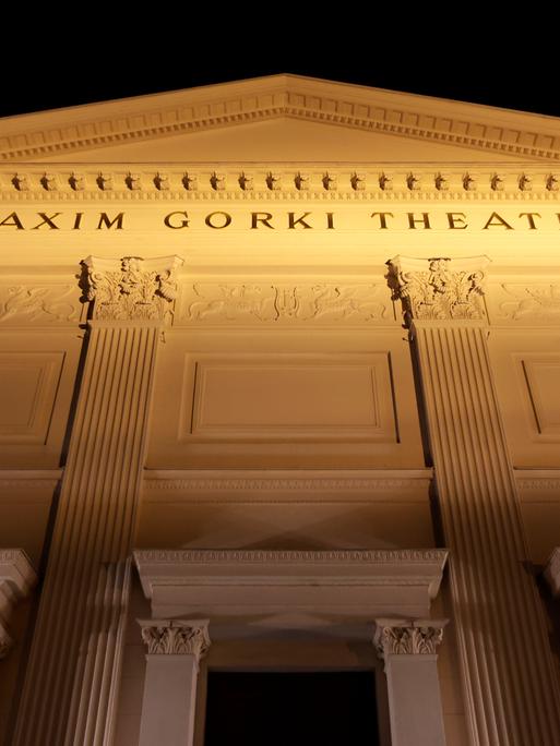 Die beleuchtete Fassade des Maxim Gorki Theaters mit klassizistischen Säulen und der Aufschrift "Maxim Gorki Theater" von unten fotografiert vor schwarzem Nachthimmel.