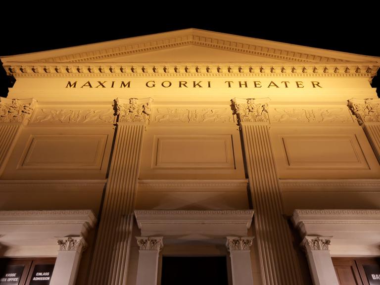 Die beleuchtete Fassade des Maxim Gorki Theaters mit klassizistischen Säulen und der Aufschrift "Maxim Gorki Theater" von unten fotografiert vor schwarzem Nachthimmel.