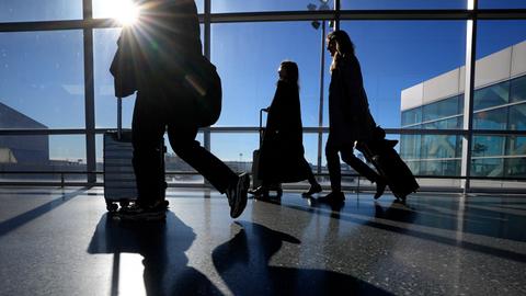 Reise schieben ihre Koffer in einem Terminal des Flughafen Boston Logan. Durch die Schreiben des Gebäudes scheint die Sonne herein.