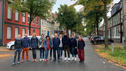 Zehn Menschen stehen nebeneinander mitten auf der Straße und schauen in die Kamera. Im Hintergrund ist eine Straße zu sehen, an deren Rändern Autos geparkt sind und Linden stehen.