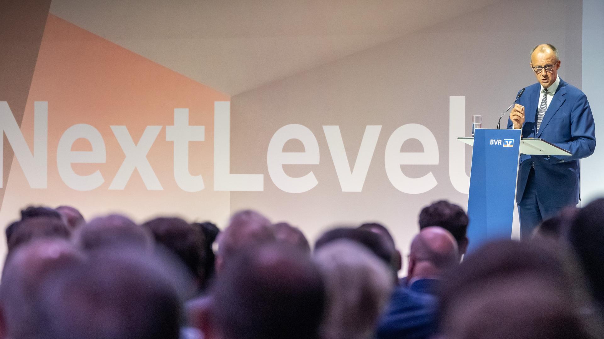 Merz steht am rednepult und spricht. Davor die Köpfe von Zuhörern. Hinter Merz eine Wand in grau-beige-Tönen mit der Aufschrift "NextLevel".