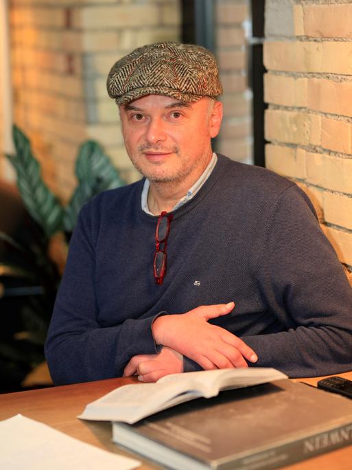 Der Schriftsteller Catalin Dorian Florescu sitzt an einem Tisch und trägt eine Schiebermütze, vor ihm mehrere Bücher.