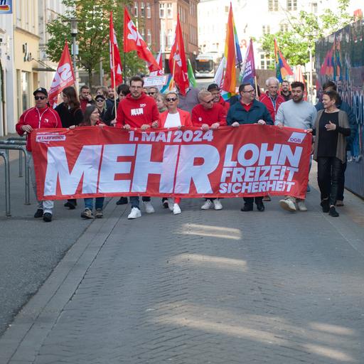 Demonstranten tragen ein großes, rotes Banner vor sich. Darauf steht: "1. Mai 2024. Mehr Lohn, Freizeit, Sicherheit"