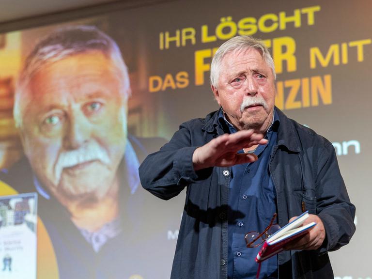 Ein Mann, der Liedermacher und Lyriker Wolf Biermann, steht vor einer Projektion seines eigenen Porträts und dem Schriftzug "Ihr löscht das Feuer mit Benzin"