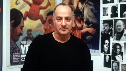 Peter Zadek sitzt vor einer Wand, die mit Fotografien und Plakaten behangen ist.