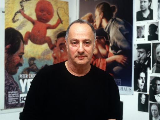 Peter Zadek sitzt vor einer Wand, die mit Fotografien und Plakaten behangen ist.