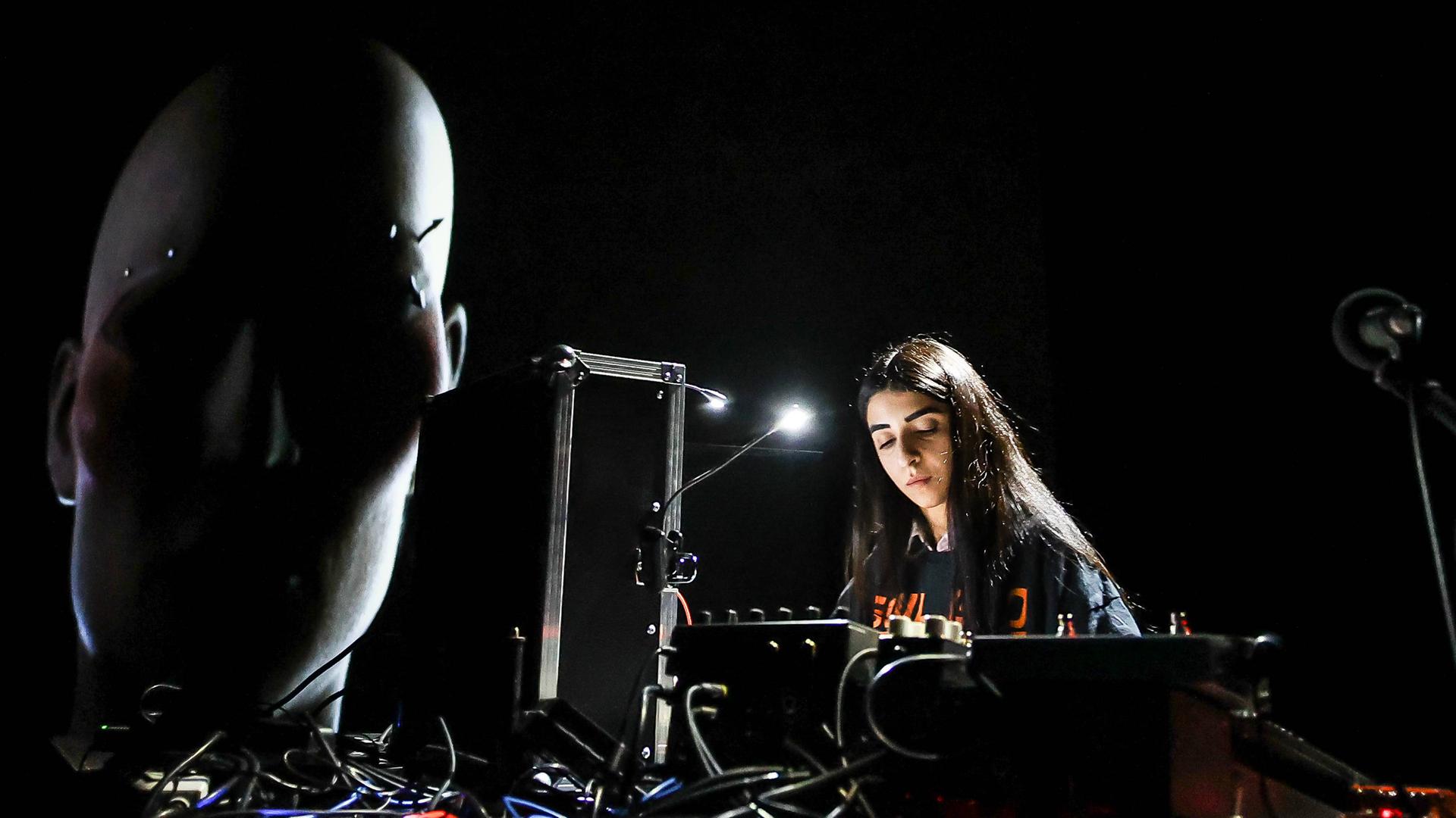 Die iranisch-deutsche Künstlerin Nazanin Noori steht bei einem Konzert beim CTM-Festival 2023 an technischen Geräten. Daneben ist ein projiziertes Gesicht zu sehen, ein Werk der Künstlerin Diana Azzuz, die einen ukrainisch-syrischen Hintergrund hat. 