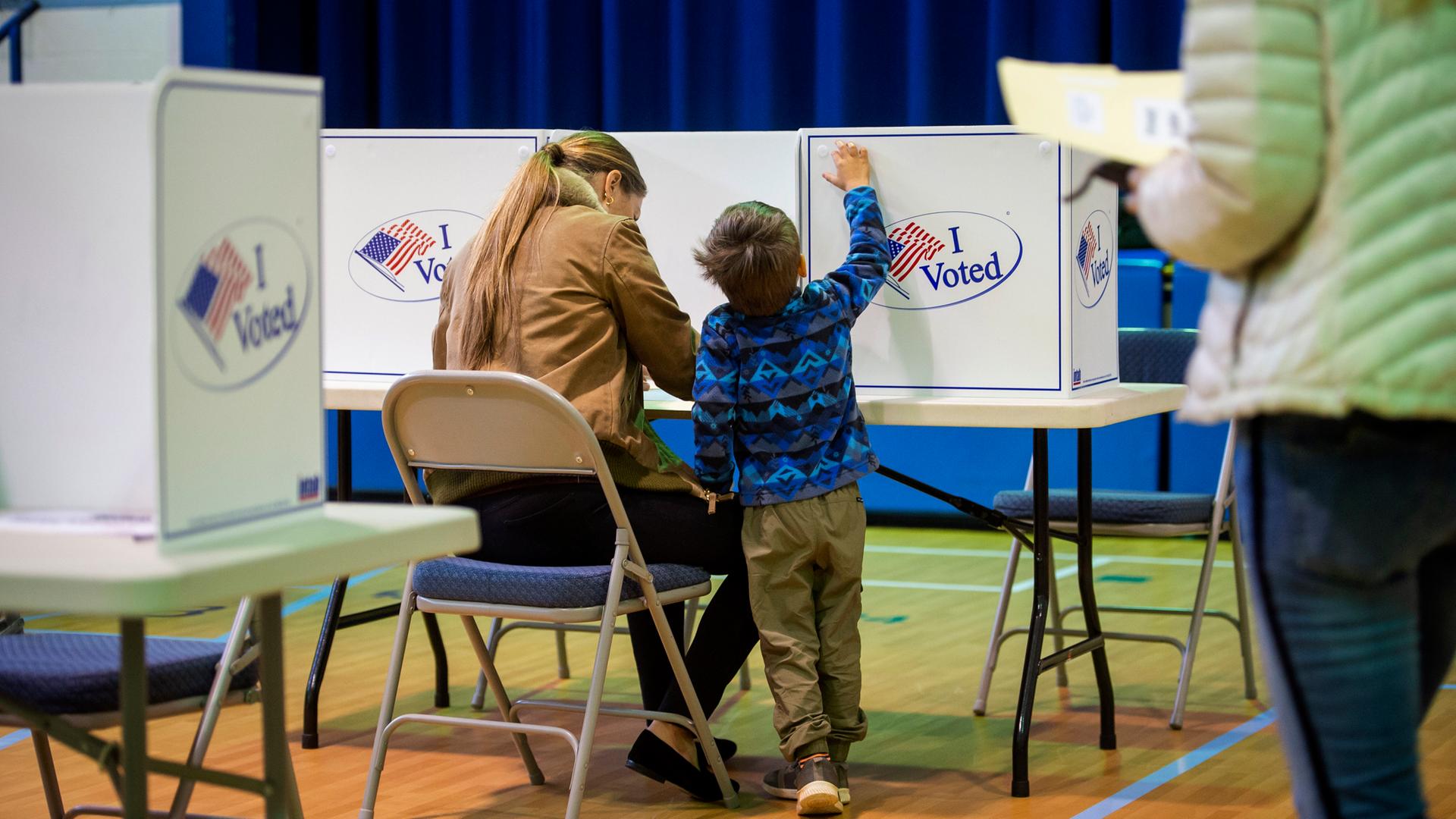 Eine Frau wählt in den USA. Sie ist von hinten in der Wahl-Kabine zu sehen, neben ihr ein kleines Kind. 