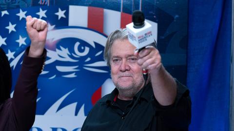 Steve Bannon hält ein Mikrofon hoch während seiner Radiosendung auf der Conservative Political Action Conference, CPAC 2023. Im Hintergrund eine Flagge der USA mit Adler.
