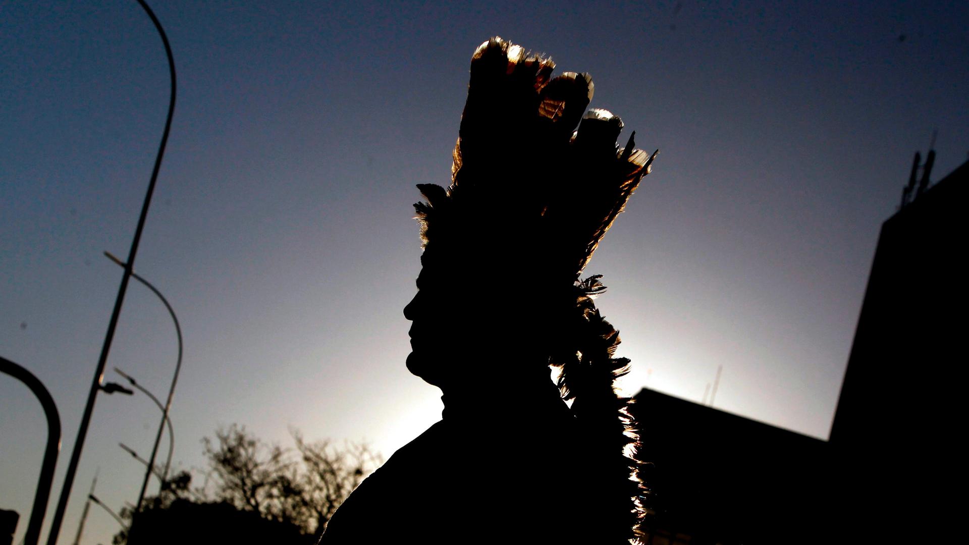 Die Silhouette eines Indigenen mit einem Federkopfschmuck hebt sich vor einem dämmerigen Himmel ab.