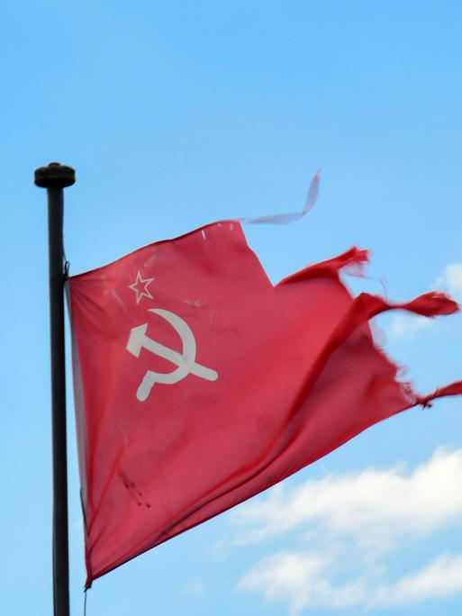 Vor blauem Himmel hängt eine zerrissene sowjetische Flagge mit Hammer, Sichel und Stern.