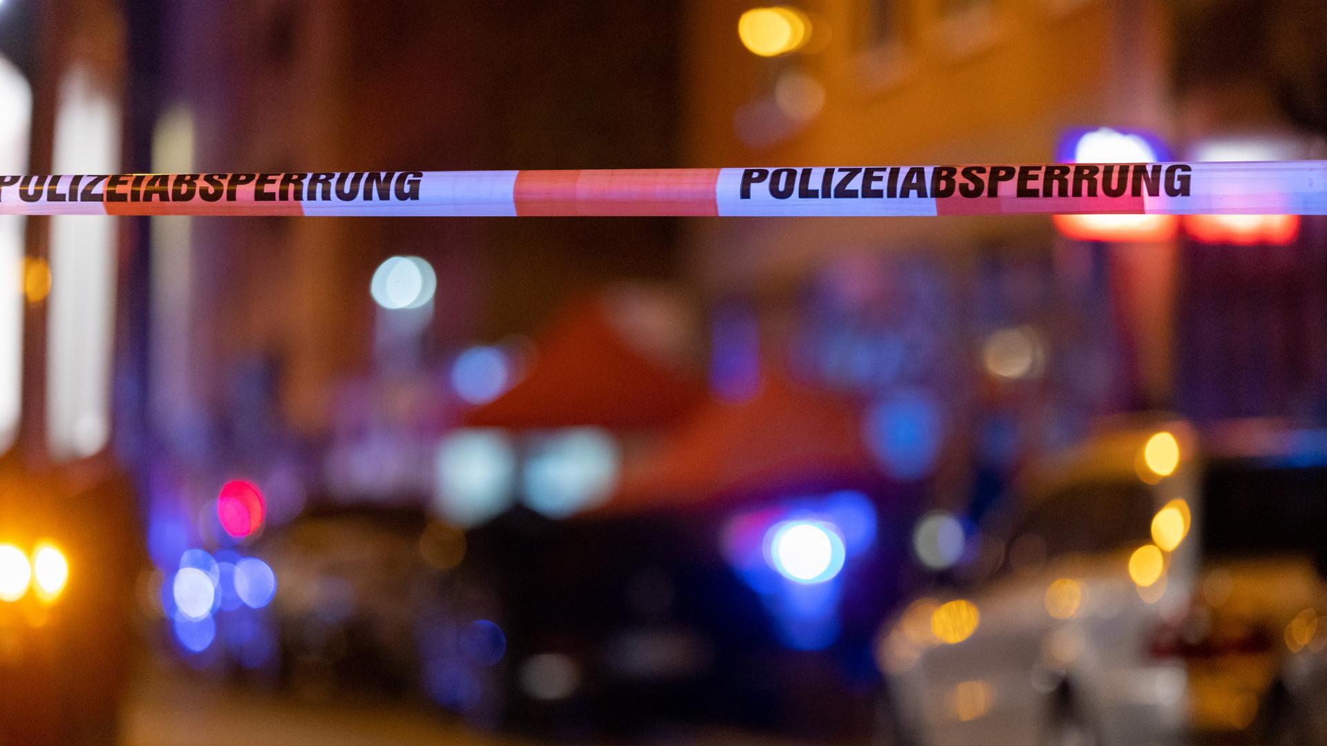 Ein Absperrband der Polizei hängt vor einem Tatort, nachdem hier Schüsse gefallen waren. Im Hintergrund stehen Polizisten neben einer Straßenbahn.
