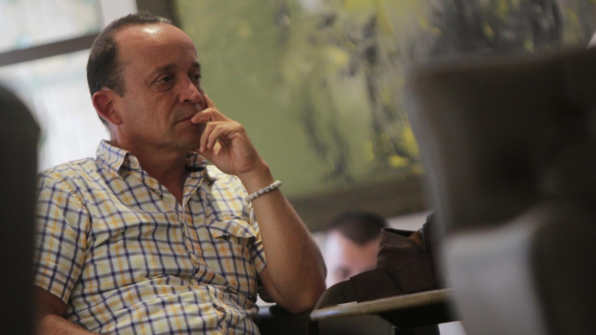 Uribe im karierten weiß-gelben Hemd sitzt in einem Raum und hält die Hand ans Kinn. Dahinter unscharf der Kopf einer weiteren Person. Uribe im karierten weiß-gelben Hemd sitzt in einem Raum und hält die Hand ans Kinn. Dahinter unscharf der Kopf einer weiteren Person.