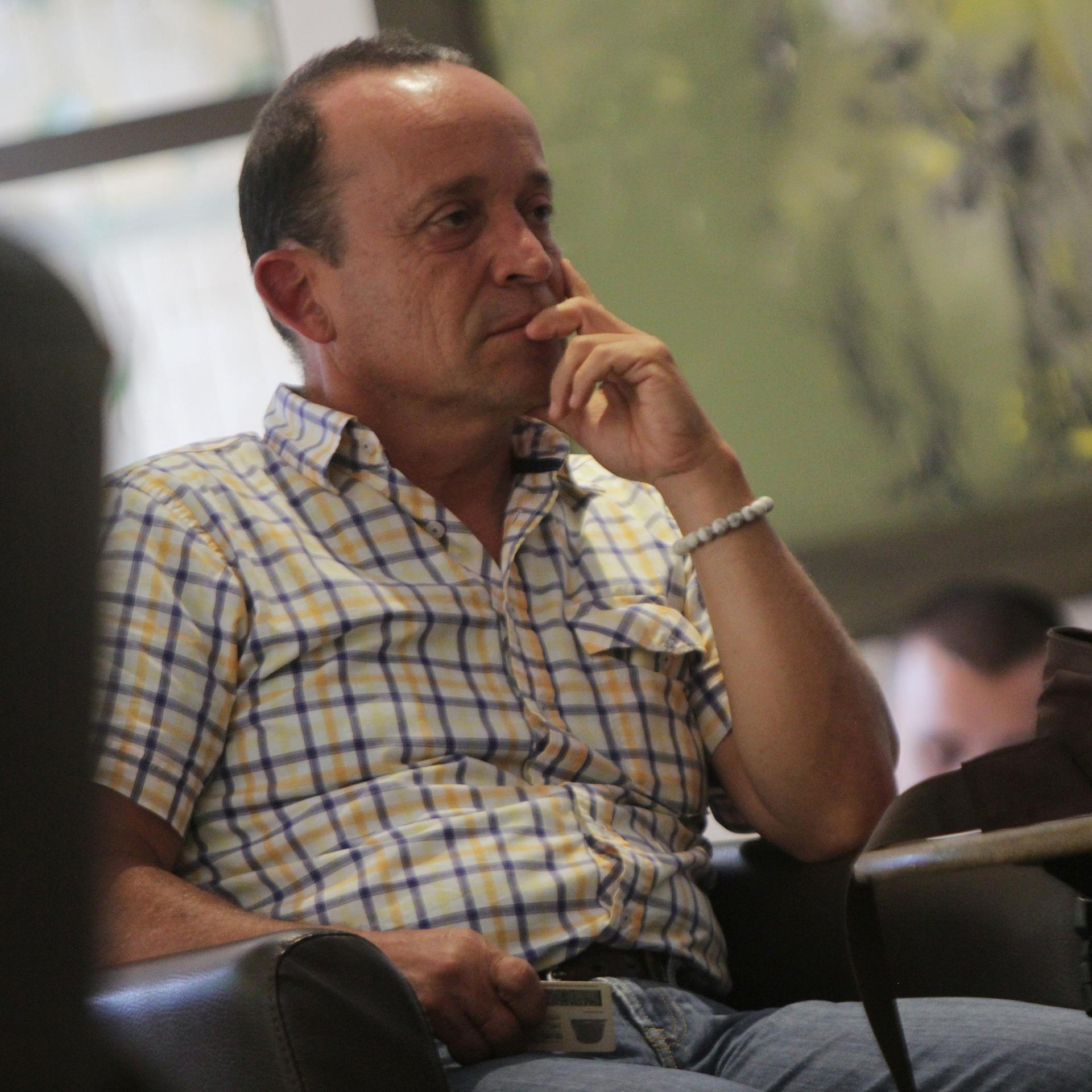 Uribe im karierten weiß-gelben Hemd sitzt in einem Raum und hält die Hand ans Kinn. Dahinter unscharf der Kopf einer weiteren Person. 
