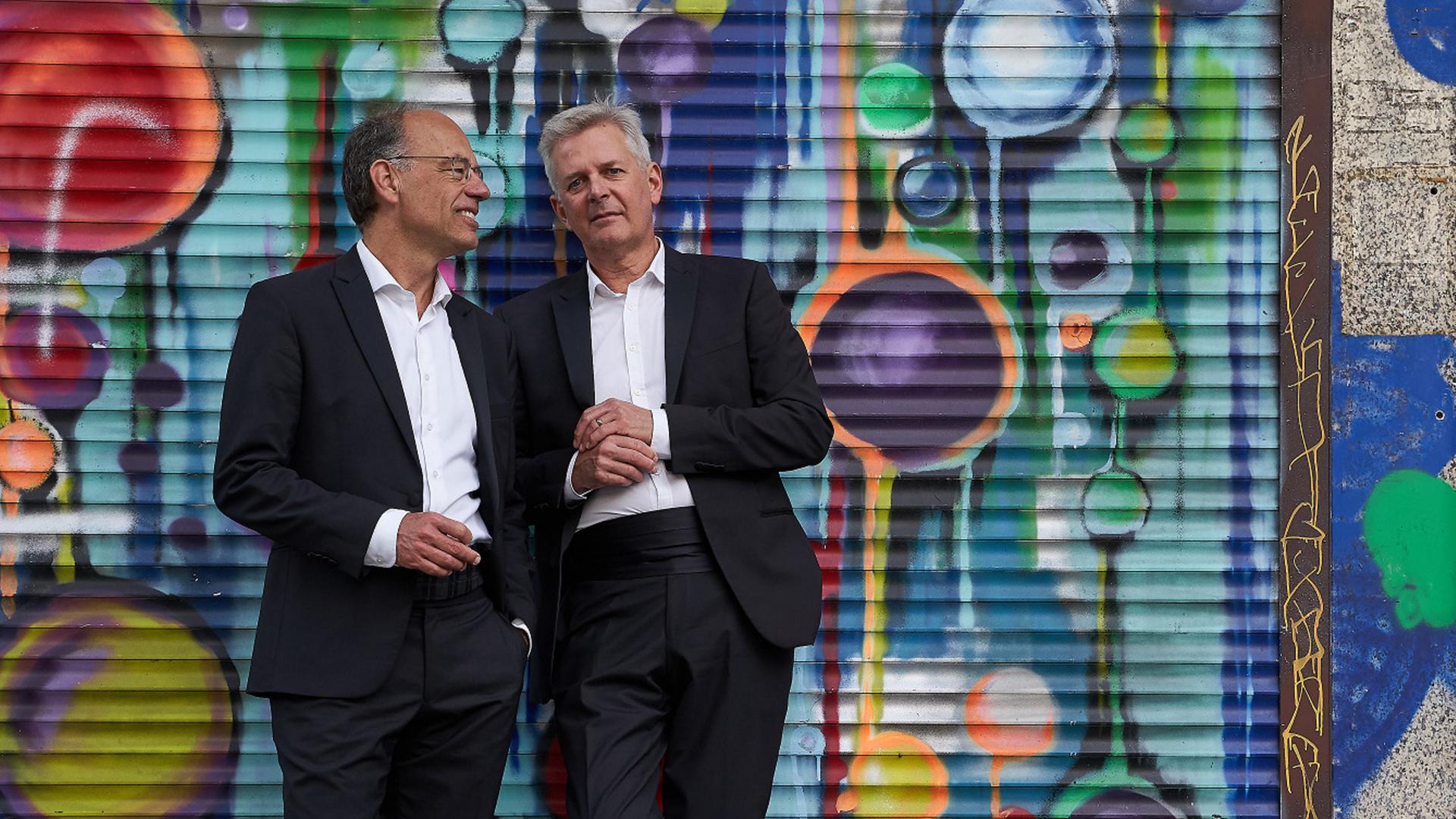 Zwei Männer stehen in Anzügen vor einer Wand, die von Sprayerns bunt gestaltet wurde. Zwei Männer stehen in Anzügen vor einer Wand, die von Sprayerns bunt gestaltet wurde.