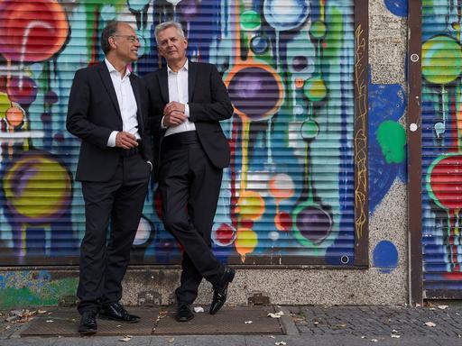 Zwei Männer stehen in Anzügen vor einer Wand, die von Sprayerns bunt gestaltet wurde. Zwei Männer stehen in Anzügen vor einer Wand, die von Sprayerns bunt gestaltet wurde.