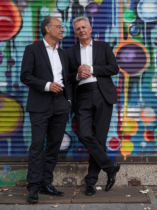 Zwei Männer stehen in Anzügen vor einer Wand, die von Sprayerns bunt gestaltet wurde.