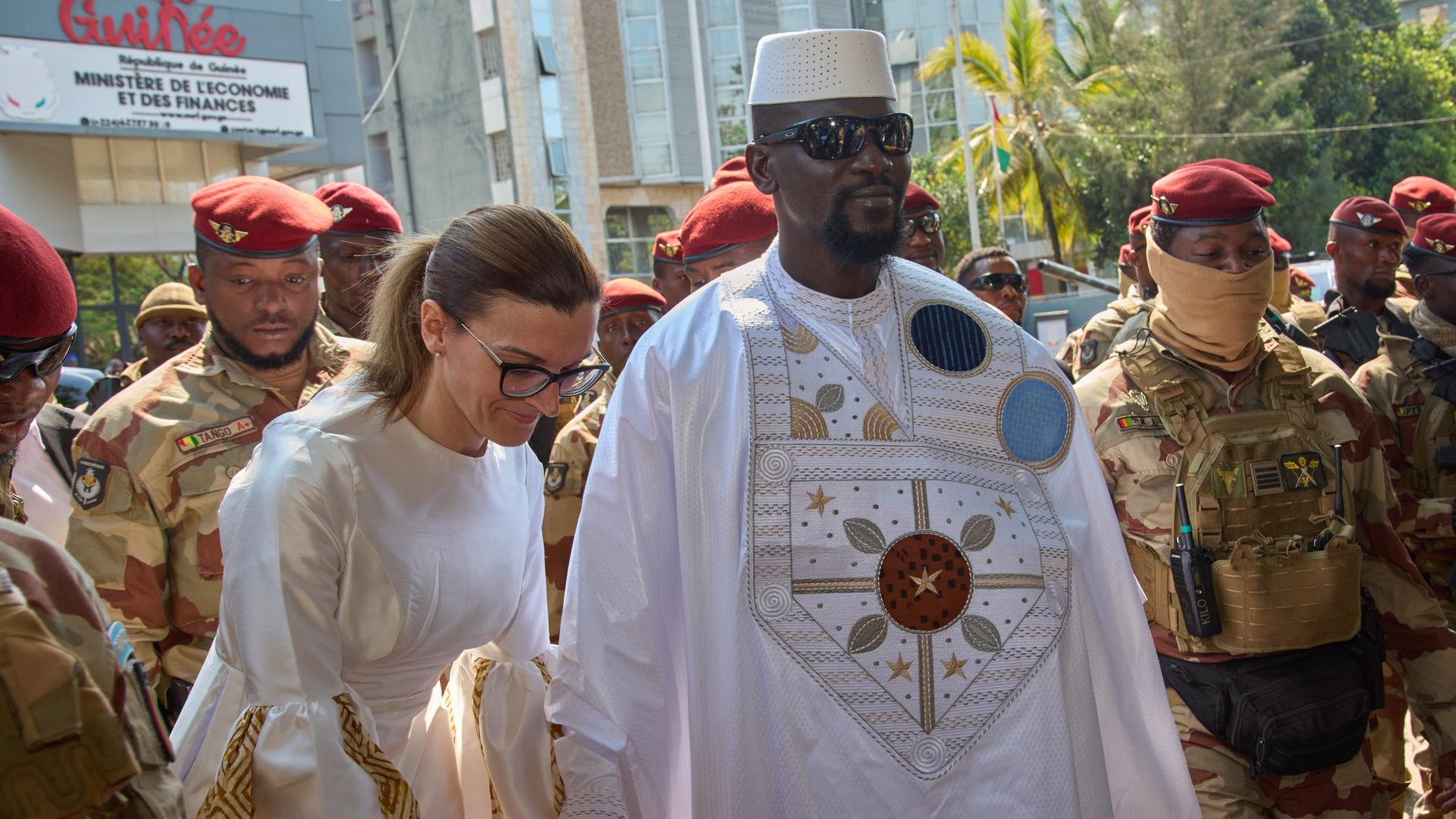 Doumbouya mit Sonnenbrille, weißem geqand udn weißer Koopfbedeckung, hält seine ebenfalls weißgekleidete weiße Frau an der Hand. Dahinter mehrere Soldaten vor Gebäuden. Doumbouya lächelt. 