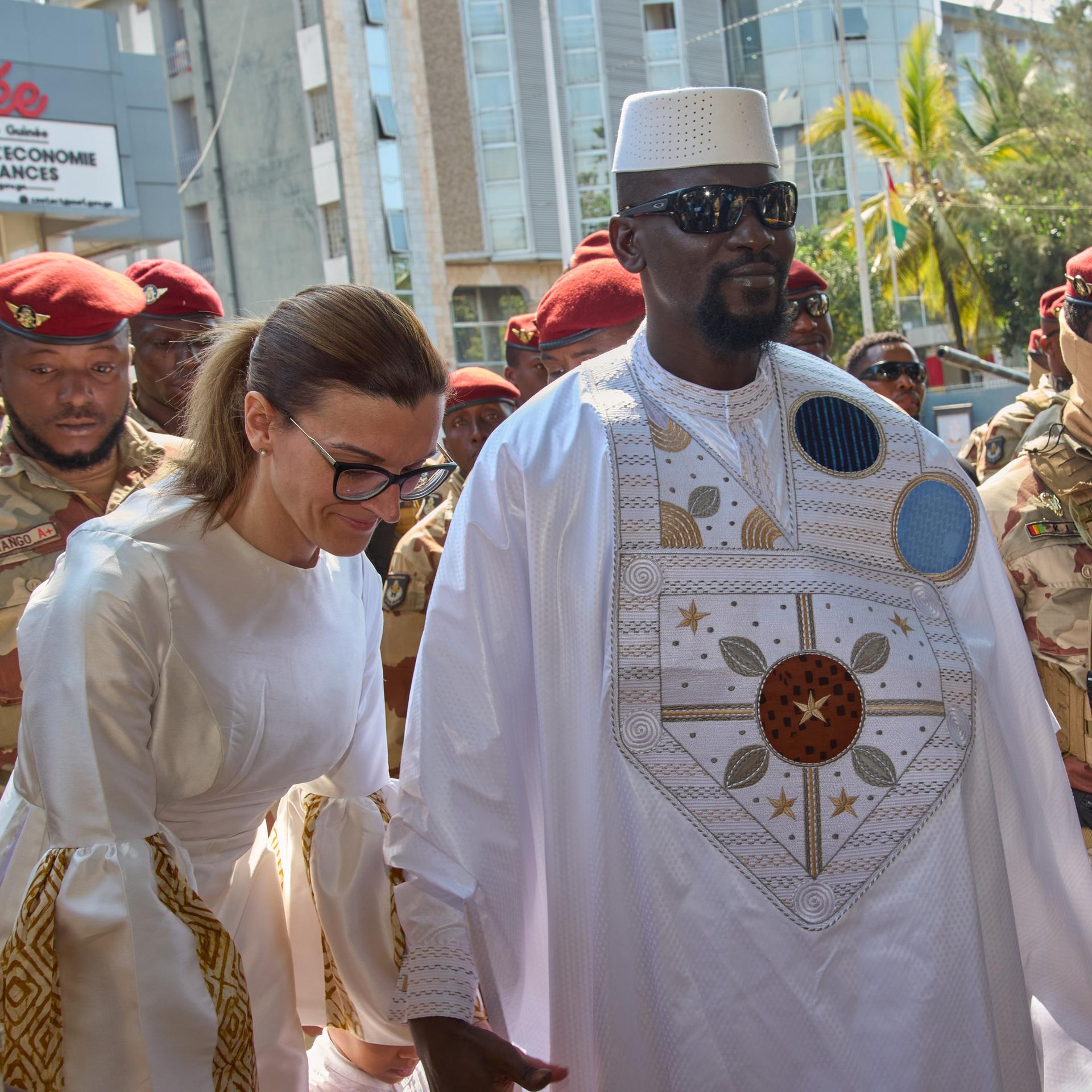 Doumbouya mit Sonnenbrille, weißem geqand udn weißer Koopfbedeckung, hält seine ebenfalls weißgekleidete weiße Frau an der Hand. Dahinter mehrere Soldaten vor Gebäuden. Doumbouya lächelt. 