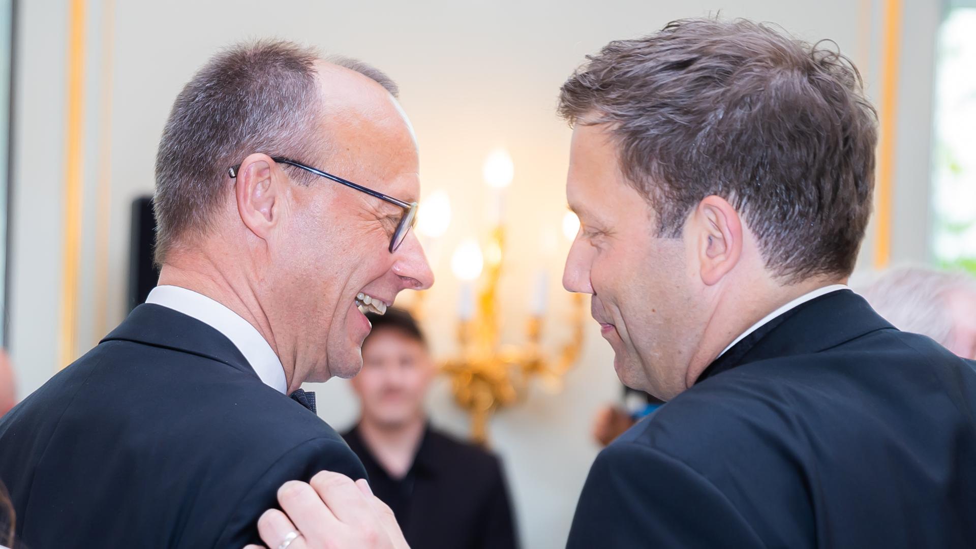 Zwei Männer in Anzügen stehen nebeneinander und schauen sich an. Der rechte Mann fasst den linken Mann an der Schulter an. Der linke Mann lacht. Es sind Friedrich Merz und Lars Klingbeil.