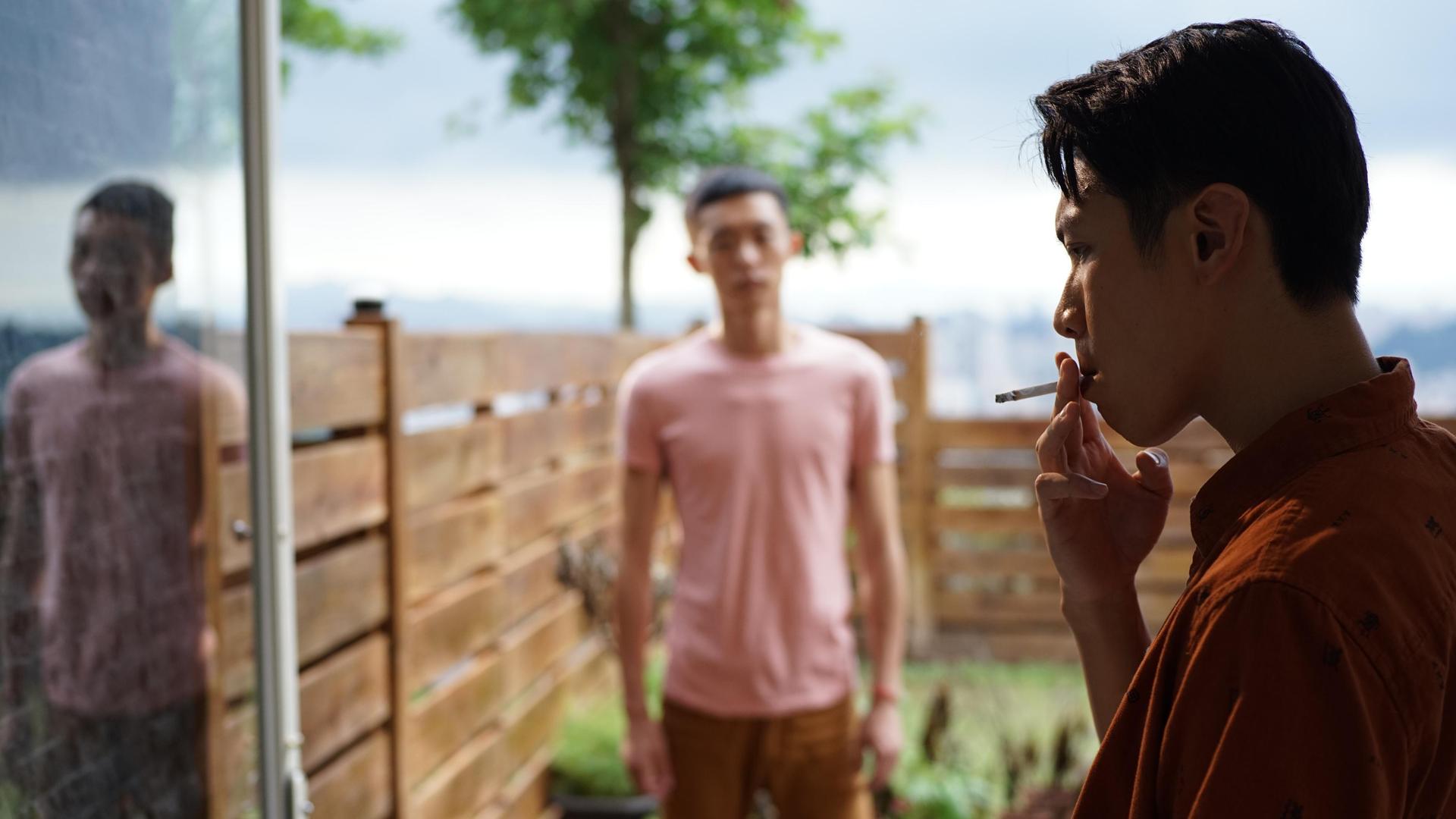 Ein Mann lehnt an einem Türrahmen und zieht cool an einer Zigarette. Im Garten dahinter steht ein weiterer Mann im Unschärfebereich des Bildes und schaut den rauchenden Mann an.