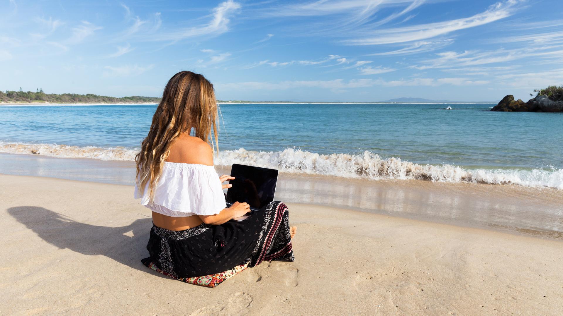 Eine junge Frau sitzt am allein Strand und bedient ein Laptop. Im Hintergrund ist das Meer zu sehen und ein paar Felsformationen im Wasser